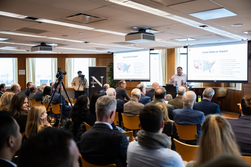 A presenter in business casual attire stands at the front of the room next to two large projection screens displaying slides titled "From Data to Impact" with maps of the United States. The audience of approximately 40-50 attendees sits in rows of wooden chairs, dressed in business attire.