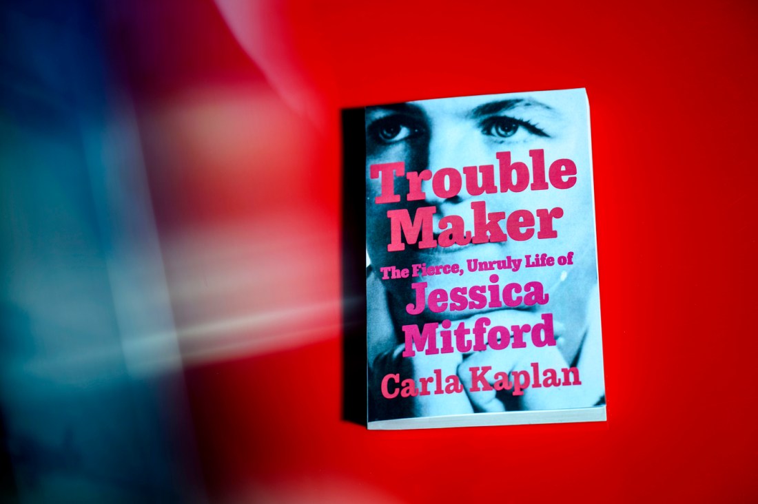 Book cover of 'Trouble Maker: The Fierce, Unruly Life of Jessica Mitford' by Carla Kaplan, featuring a close-up black and white photograph of eyes against a blurred red and blue background.