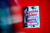 Book cover of &#039;Trouble Maker: The Fierce, Unruly Life of Jessica Mitford&#039; by Carla Kaplan, featuring a close-up black and white photograph of eyes against a blurred red and blue background.