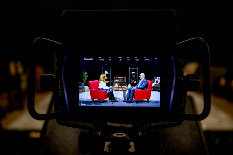 View through a camera viewfinder showing two people seated in red chairs having a conversation in a well-lit, homey studio setting with decorative shelving in the background.