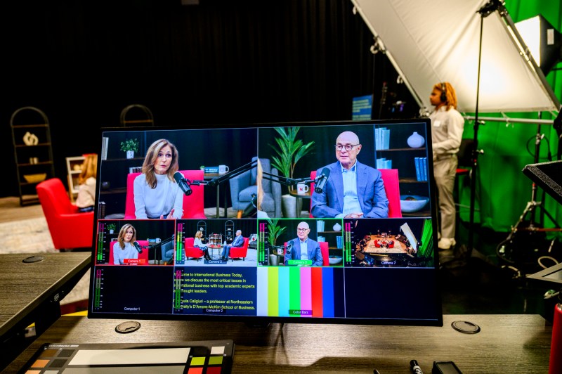 Behind-the-scenes view of a video monitor displaying a multi-camera production interface showing two people in conversation in a studio with red chairs, with production equipment and studio lighting visible in the background.