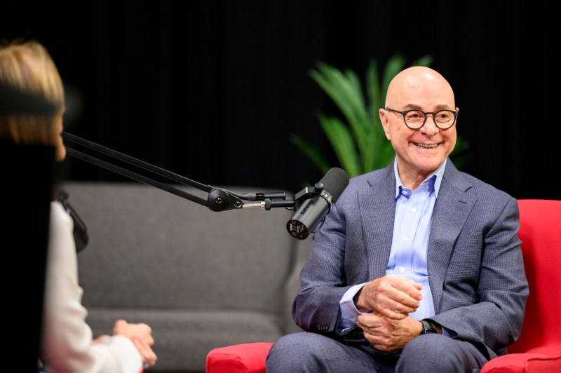 A man with glasses, wearing a grey suit, smiles while sitting in a red chair.