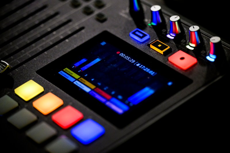 Close-up of an electronic music device or synthesizer with a dark interface displaying blue waveforms on screen, surrounded by colorful buttons in yellow, orange, red, and blue, with additional control knobs and sliders visible at the top