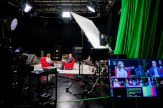 Wide view of a studio with two people in red chairs being filmed, surrounded by cameras, lighting equipment with production monitors in the foreground.