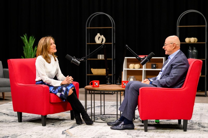 A woman in a white long-sleeve shirt and jeans talks to a man in a grey suit as they sit across from each other in large red chairs. Two podcast microphones on stands are in front of each of them.