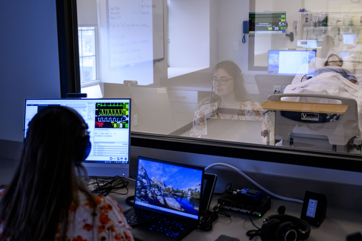 A simulation room in a medical training facility showing a student or healthcare professional at a computer workstation monitoring a medical training mannequin in an adjacent room. The observer, wearing glasses and a floral print top, sits at dual monitors displaying vital signs and medical data with green waveform patterns. 