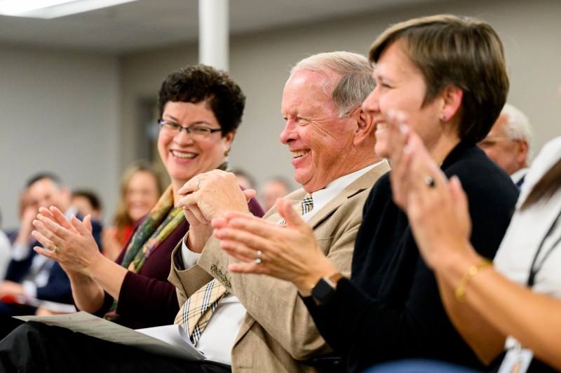 A group of people including a woman, man in a tan suite, and a woman in a black suite jacket applaud.