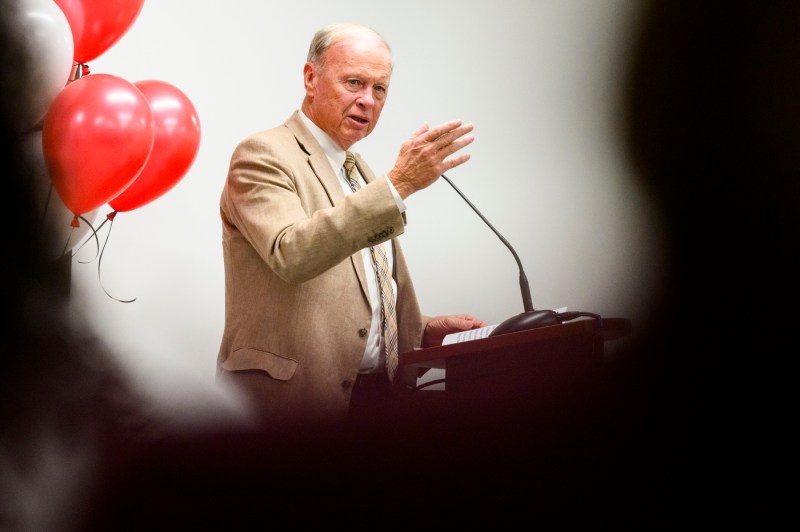 A man in a tan suit stands at a podium with his arm raised, gesturing as he speaks. Red and white balloons are seen in the background.