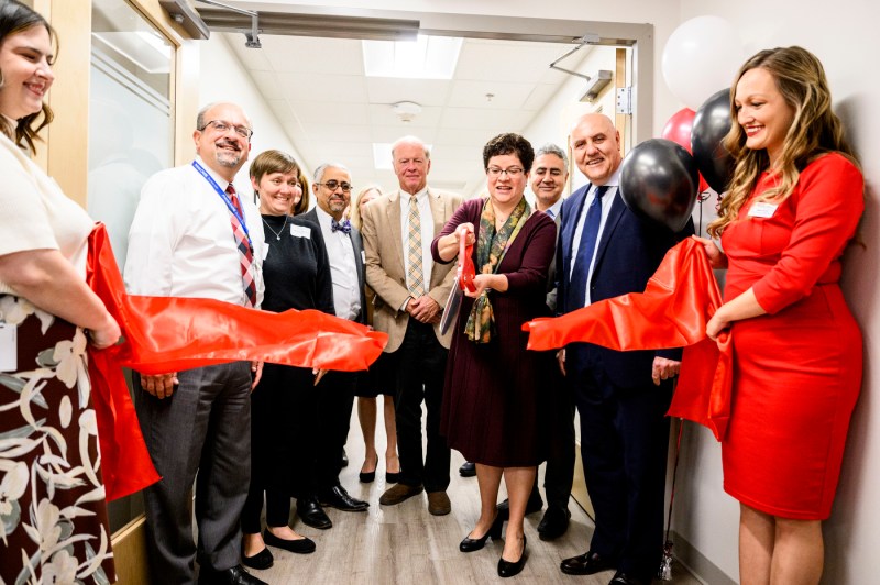 A ribbon-cutting ceremony fir the grand opening of Prima Care taking place in a bright hallway with skylights. A group of approximately ten people stand together as two people hold large red ribbons stretched across the space. The group is formally dressed, with several men in suits and women in professional attire.