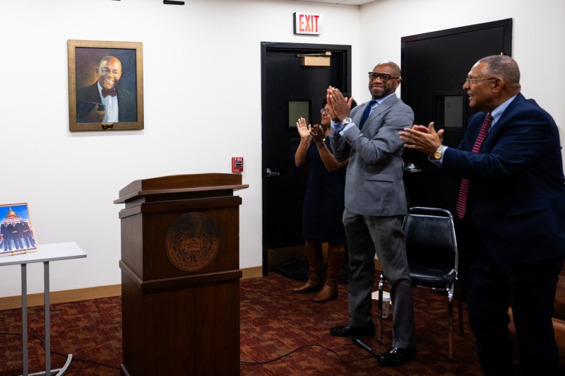 A woman and two men applaud in front of an oil painting of Northeastern alumnus William Mo Cowan.