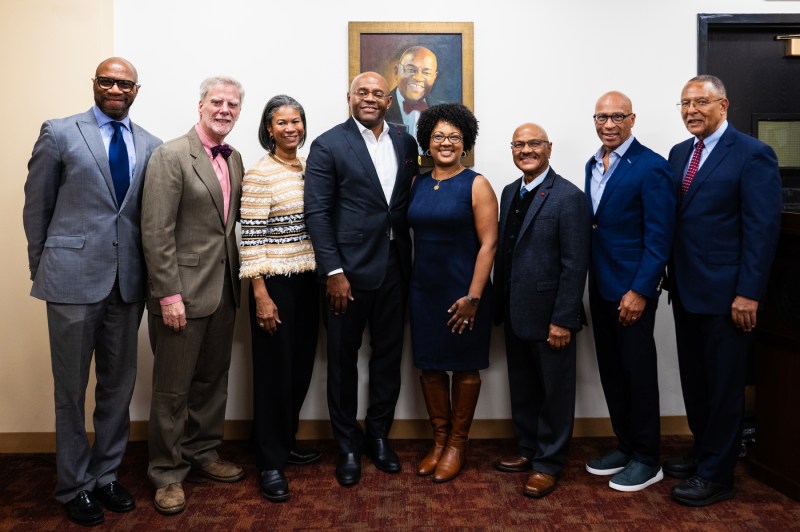 A group of eight people, male and female, dressed in business attire smile in front of an oil painting.
