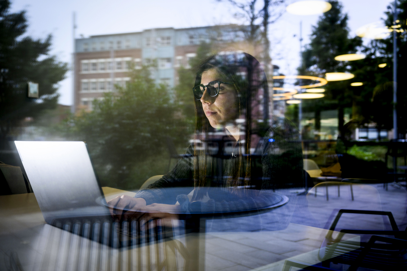 Seen through a glass window, woman in glasses and a black sweater works on a computer at a table. 