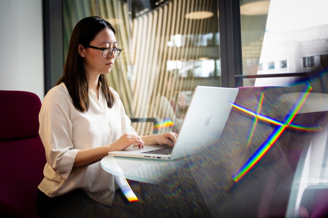 A woman in a white shirt and glasses works at her laptop against a reflective window.
