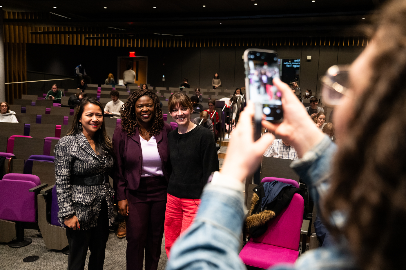 Three women stand in front of a woman pictured from behind holding a cellphone up to take a photograph. From left to right one wears a grey suit jacket, the second wears a purple jacket and the third wears a black sweater.