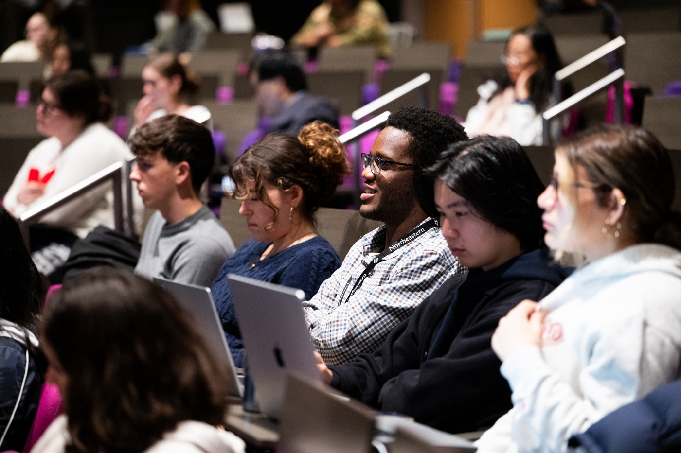 Audience members seated in an auditorium listening attentively to a presentation. In focus are several people, including a person in a checkered shirt smiling with a laptop. The tiered seating extends into the background where more attendees can be seen sitting in purple chairs.