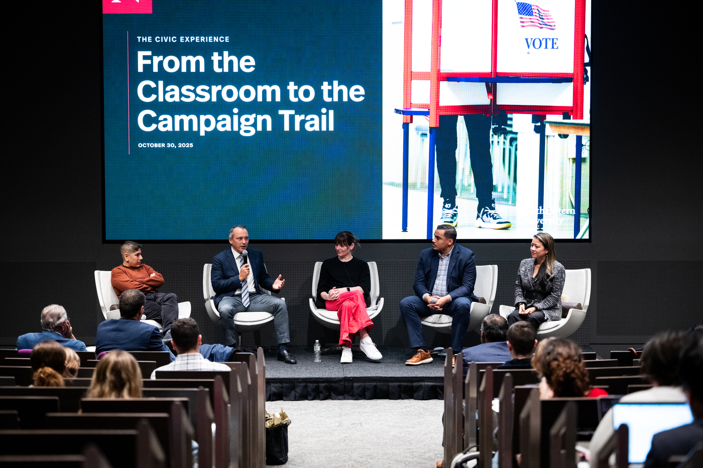 Four panelists and a moderator sit in white modern chairs on a stage before an audience, with a large screen behind them displaying the event title and an image of someone holding a 'VOTE' sign at a polling location." Panel discussion at The Civic Experience event titled 'From the Classroom to the Campaign Trail."