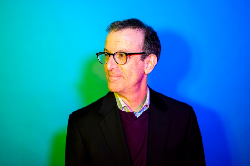 Portrait of Allen Feinstein wearing glasses posing in front of a blue gradient background.