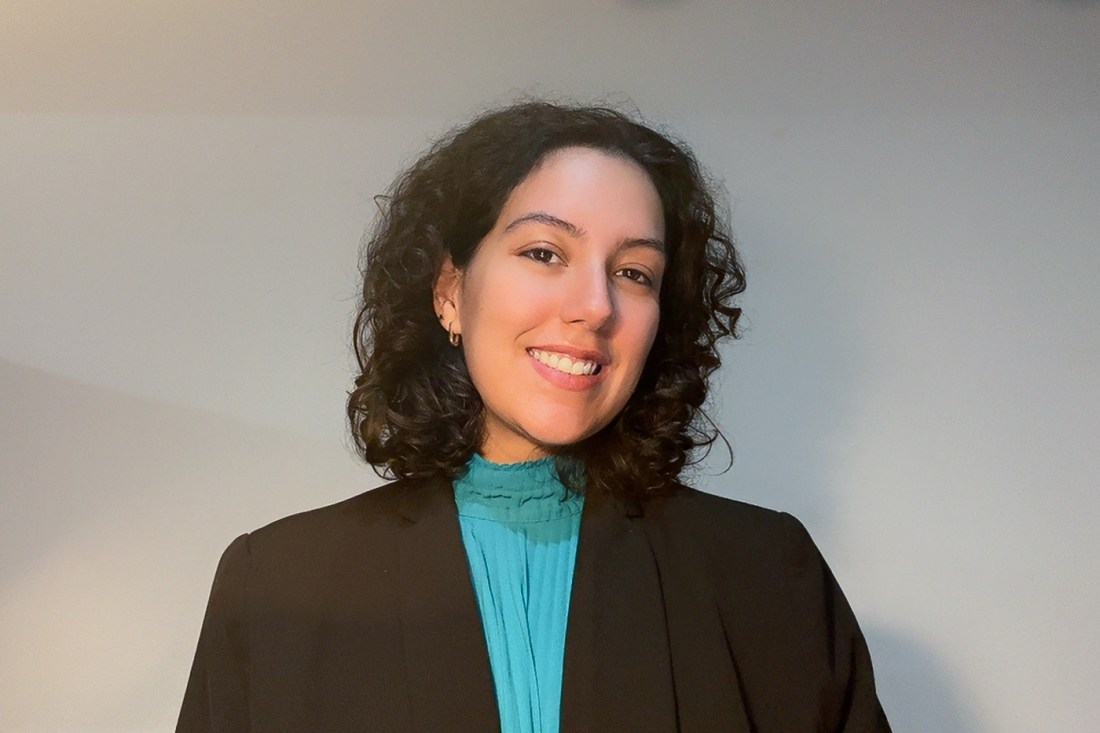 Andrea Orozco, a young woman with dark curly hair, dressed in a black blazer and turquoise top, smiles against a gray backdrop.