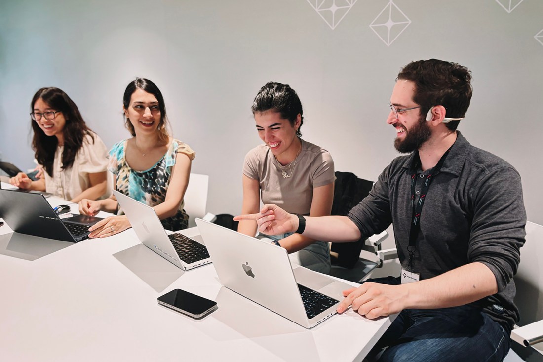 Four students, three women with dark hair and a man with glasses, in conversation sit together at a desk.