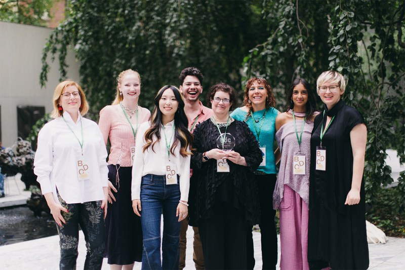 Group photo of seven people standing outdoors, smiling at the camera, wearing name badges and lanyards, with trees and greenery in the background.