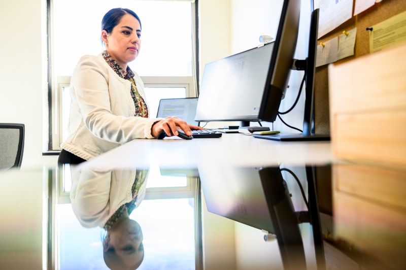A woman with dark hair pulled back and a white coat over a patterned blazer sits at a modern desk, working at a computer with several monitors. 