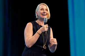 Elizabeth Gilbert, a white woman wearing a blue blouse, smiles while speaking into a microphone.