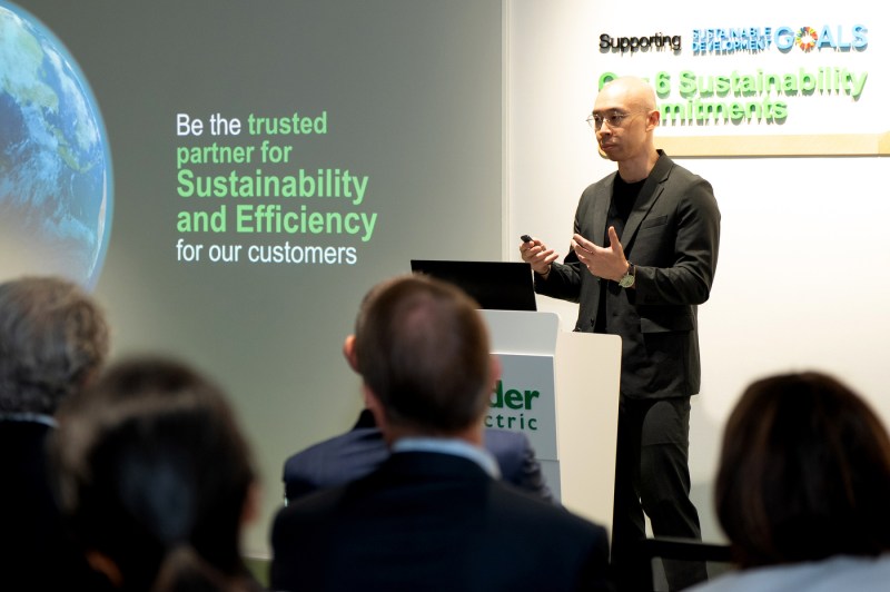 A speaker in a dark blazer stands at a podium and speaks to an audience while gesturing. Next to them, a presentation slide shows a picture of the earth with the words 'Be the trusted partner for Sustainability and Efficiency for our customers'. 