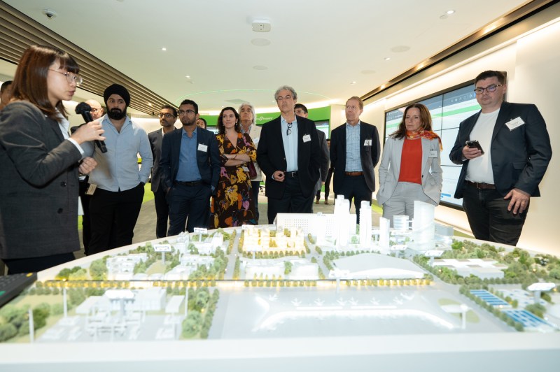 A group of people gather around an architectural scale model on a display table. A woman holding a microphone appears to lead the discussion about the development project. 