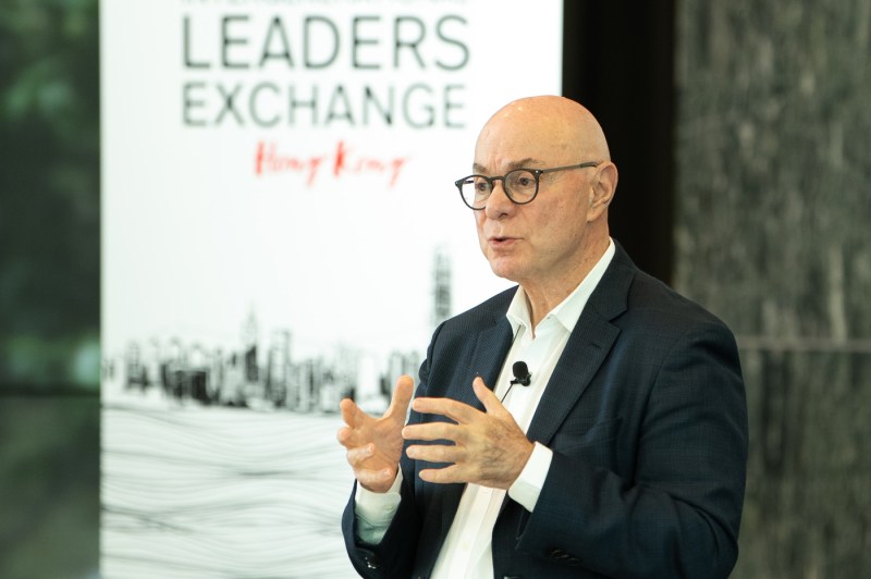 President Aoun wearing glasses, a dark blazer, and white button down shirt with a microphone attached, gestures while speaking at an event. A white banner that says 'Leaders Exchange Hong Kong' is visible in the background.