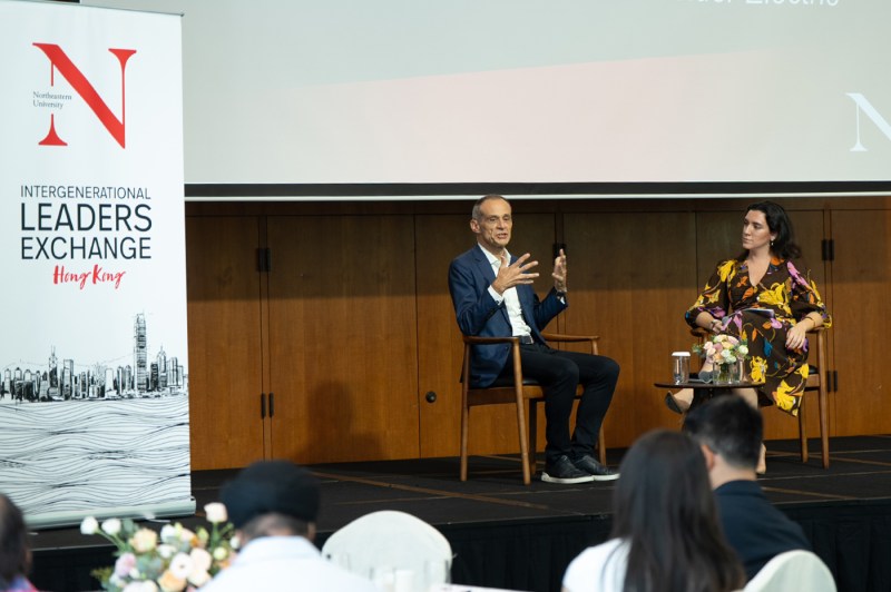 Two panelists sitting on stage in wooden chairs. One is speaking while gesturing. Next to them is a white banner that has the Northeastern logo and the words 'Intergenerational Leaders Exchange Hong Kong' on it. 