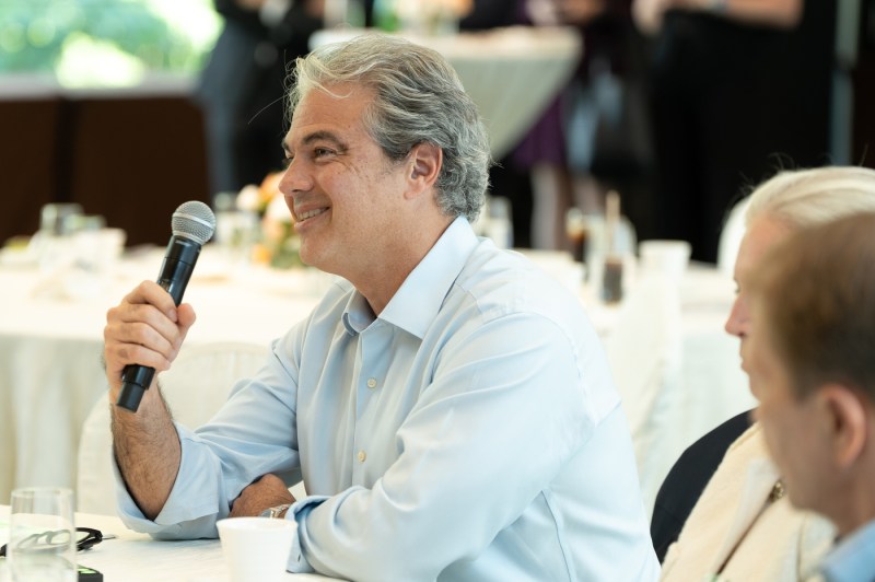 A man in a light blue shirt smiles while holding a microphone during a seated discussion. Two other attendees are visible in the foreground. 