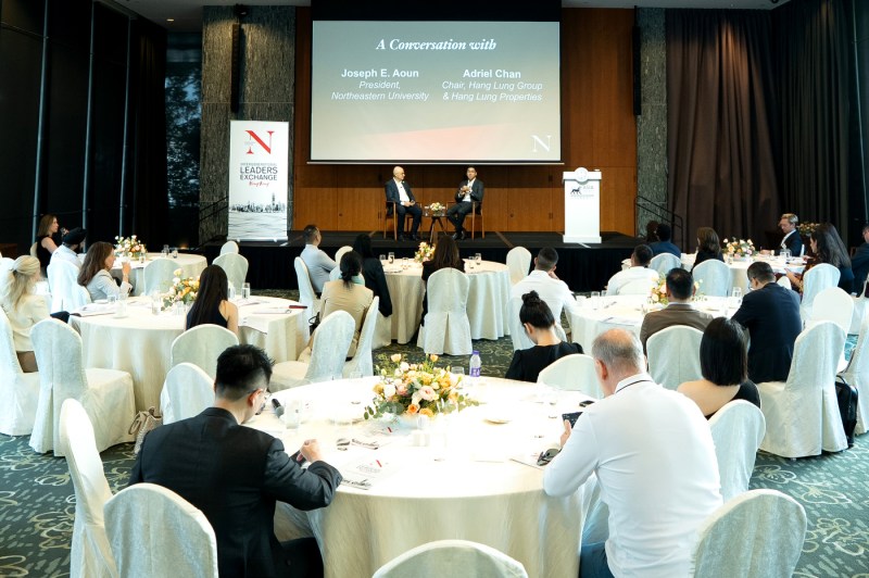 President Aoun and Adriel Chan sitting in modern wood chairs on a stage in front of a room of audience members sitting at circular tables with white table cloths.