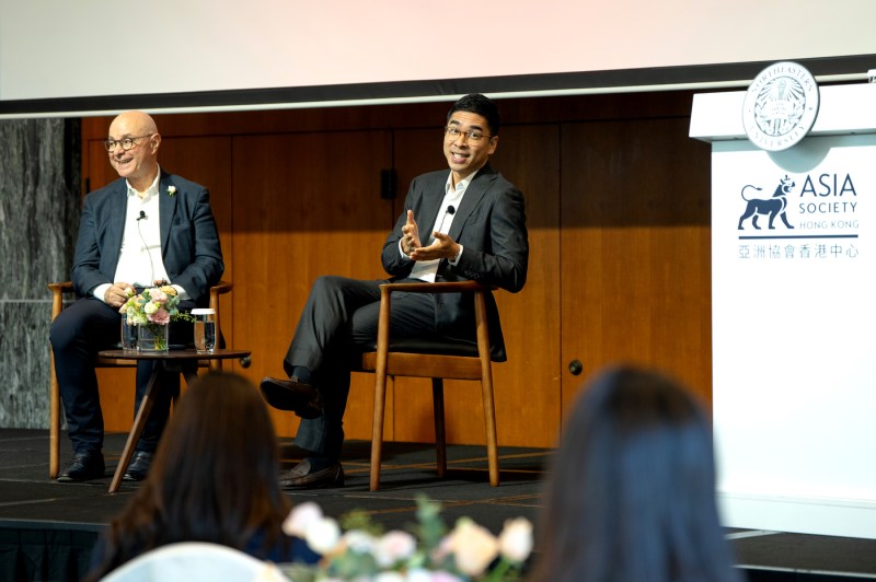 Two panelists sit in wooden chairs on stage at the 'Integenerational Leaders Exchange' event in Hong Kong. They are engaged in conversation in front of an audience. 