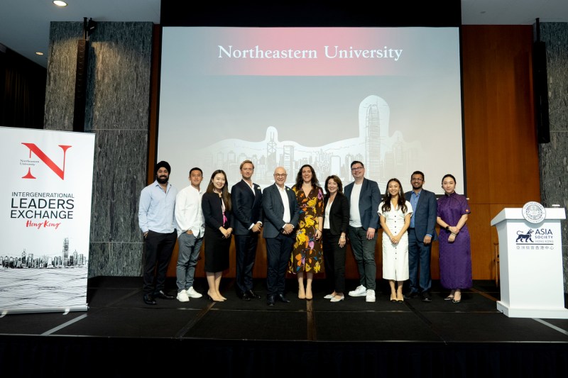 A group of approximately ten event participants and speakers pose together on stage in front of a Northeastern University branded presentation screen and 'Intergenerational Leaders Exchange Hong Kong' banner. 