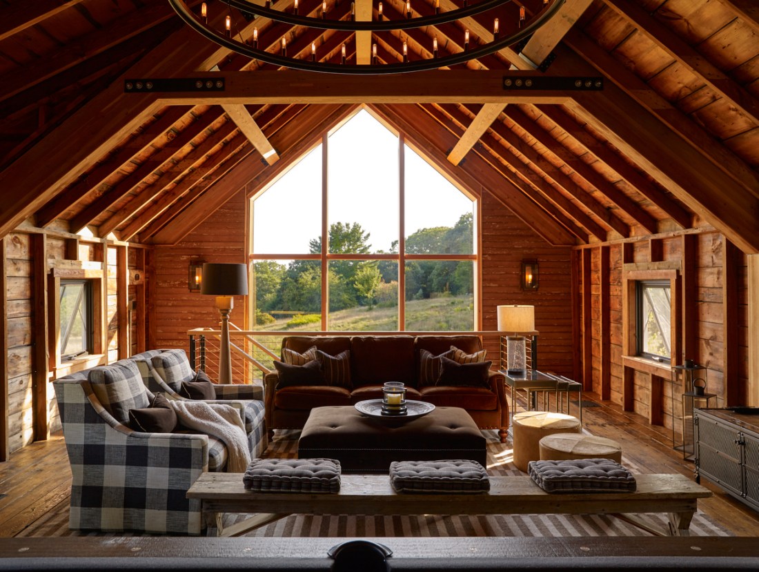 Interior of an A-frame cabin with exposed wooden beams and a cathedral ceiling. There is a seating area with plaid chairs and ottomans and a large triangular window overlooking trees.
