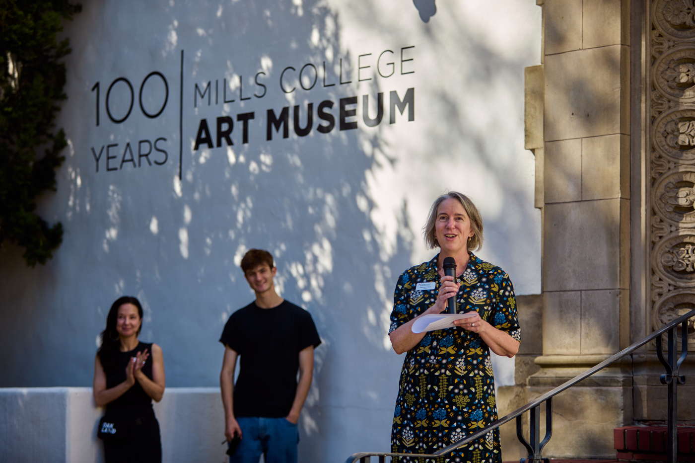 Mills College Art Museum Director Stephanie Hanor speaking into a microphone in front of wall displaying the text 'Mills College Art Museum: 100 Years.'