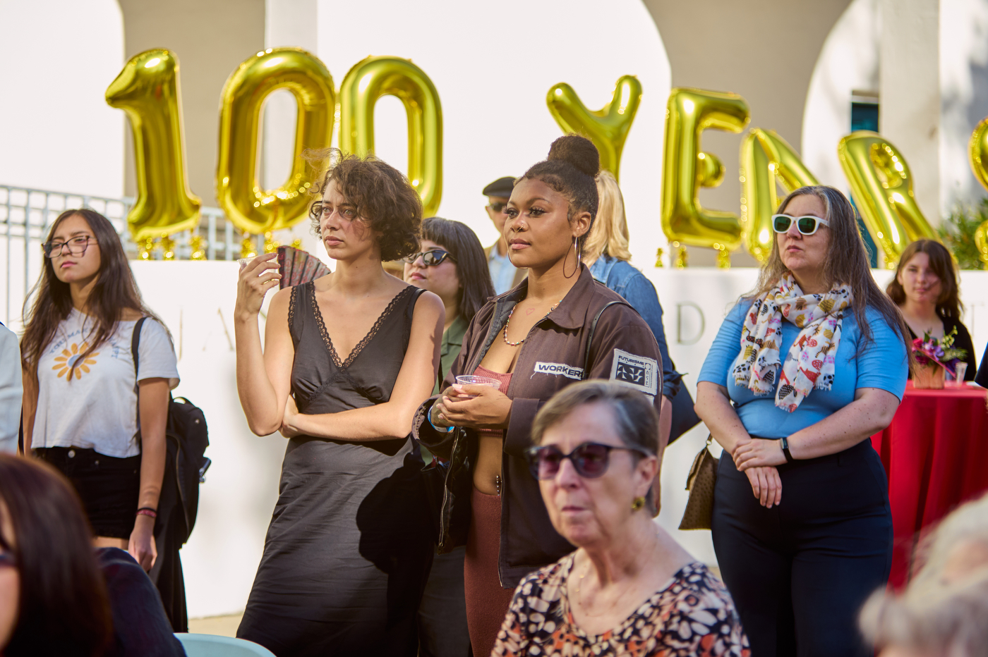 Visitors at the museum holding drinks and listening to a speaker, with yellow balloons spelling out '100 Years' in the background.