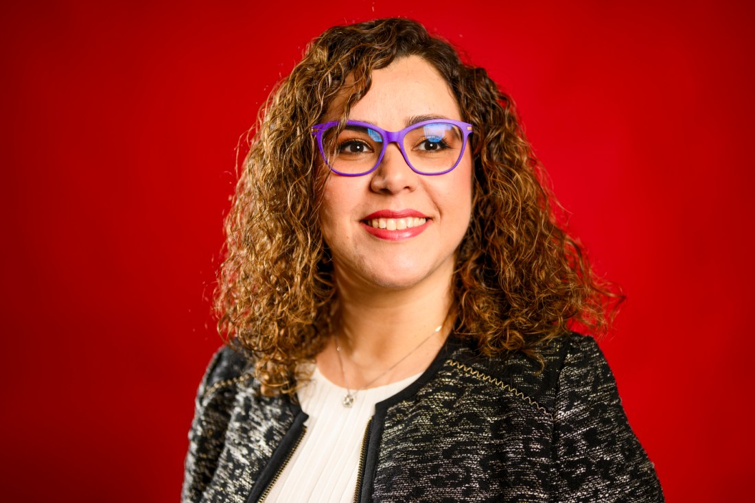 A portrait of associate professor Serena Alexander wearing purple glasses with curly hair smiling for a photo in front of a red background.