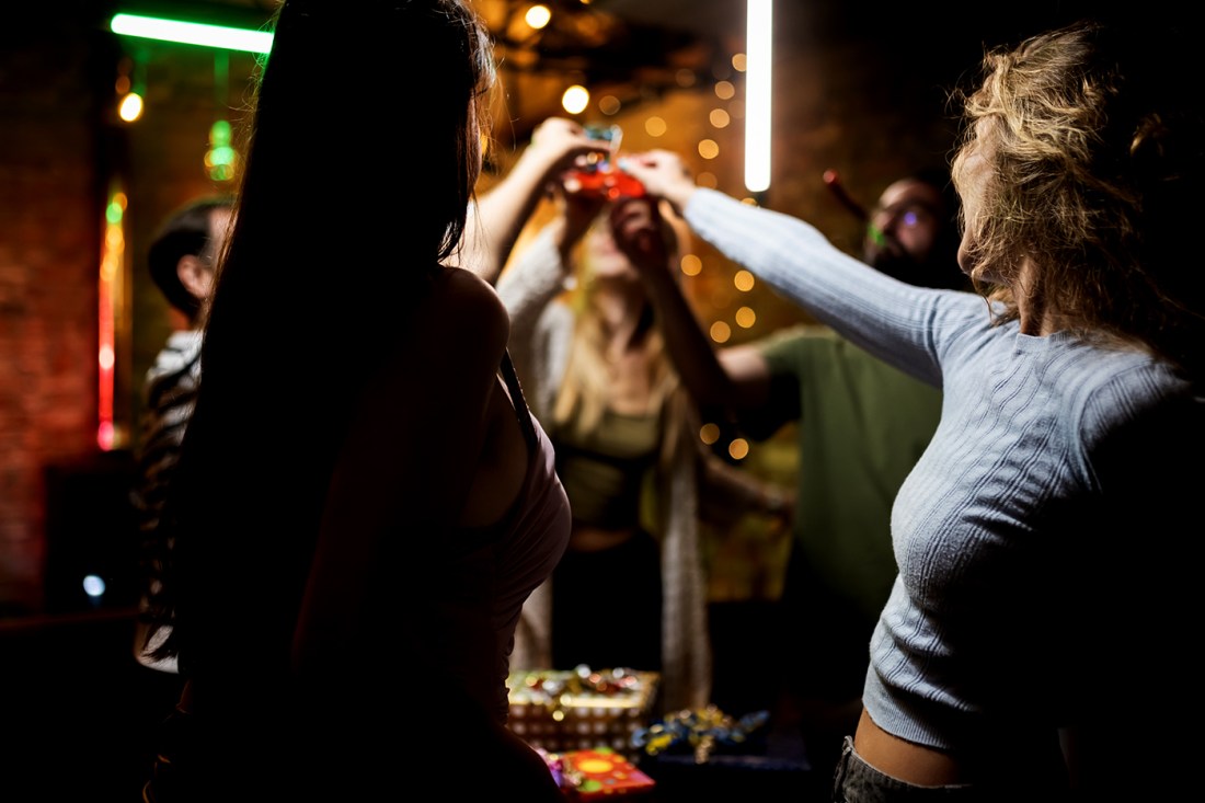 Three college-age women hold up drinks to cheers at a bar or pub.