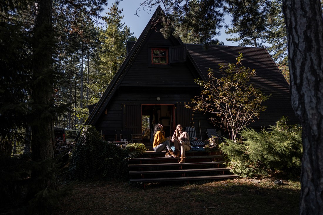 Two people sitting on the sunlit steps outside of an A-frame rental house surrounded by trees.