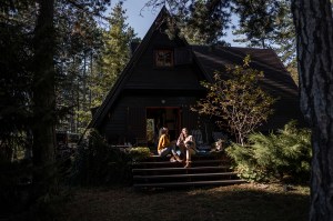 Two people sitting on the sunlit steps outside of an A-frame rental house surrounded by trees.