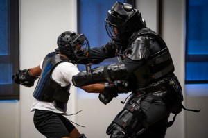 Two Black men with armor and face masks engage in a self-defense drill.