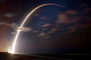 A blurry nighttime photo photo of a rocket, generating an orc of light across the night sky.