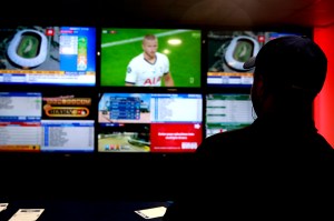 A man shrouded in shadow watches sports and engages in sports betting on a large screen.