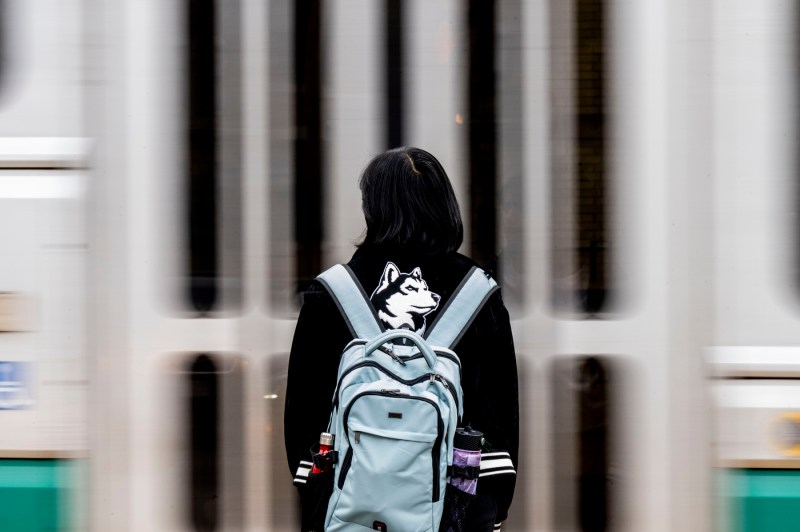 A person wearing Northeastern gear and a backpack stands in front of a green line train.