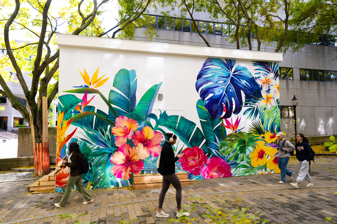 Students walk across campus next to a white building with colorful tropical flowers painted on it. 
