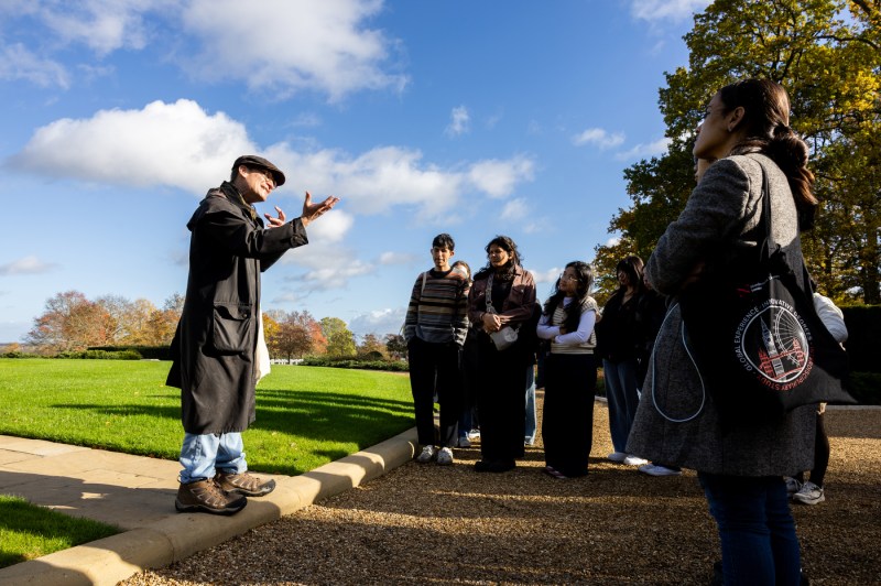 A man wearing a hat and long coat stands outside surrounded by a group as he gestures with his hands up. 
