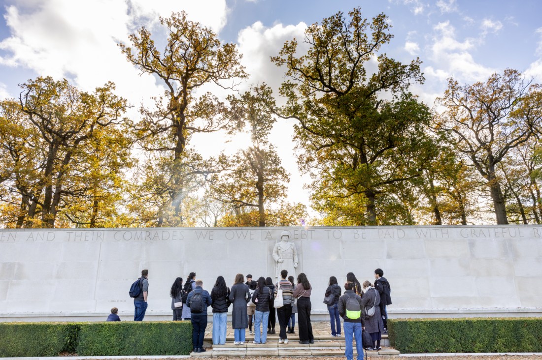 A group of people standing in front of a memorial at the Cambridge American Cemetary. In the background tall trees with autumn foliage are backlit by the sun. 
