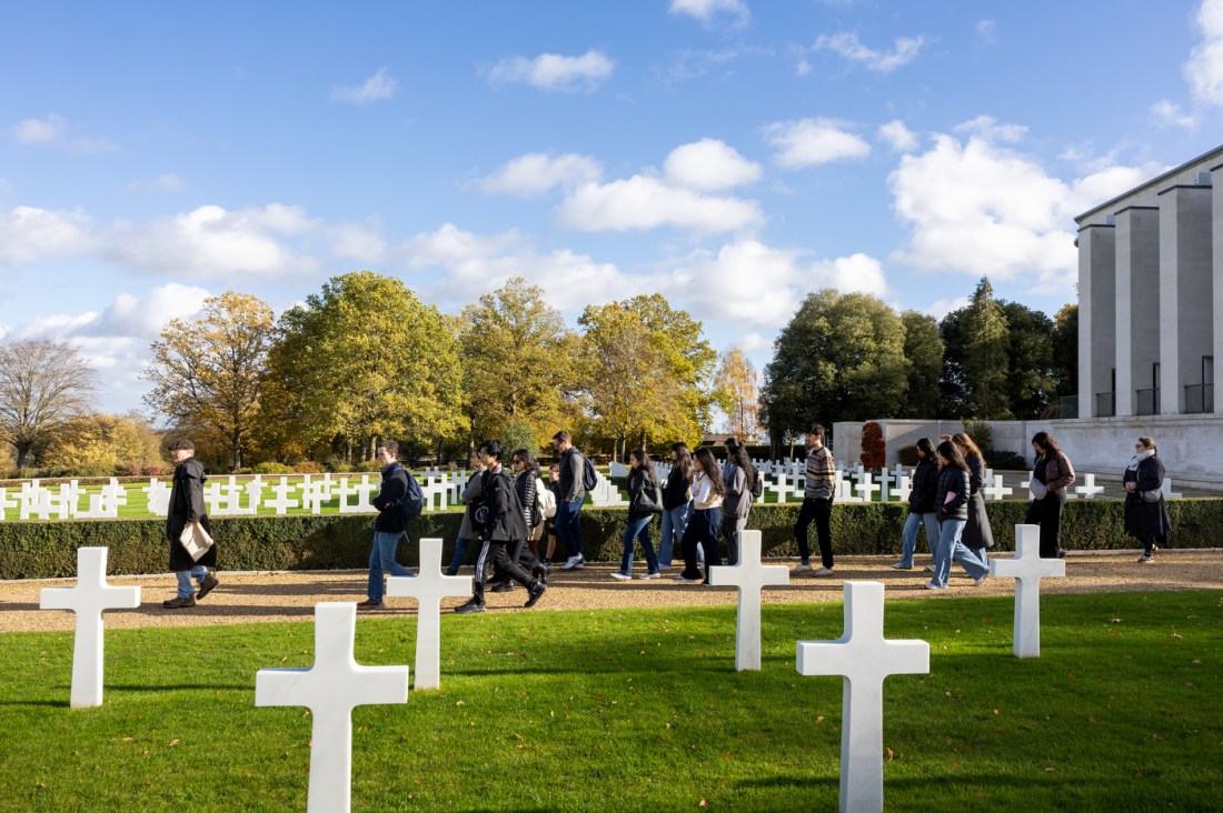 A group of people walking on a pathway next to white cross markers on a lawn at the Cambridge American Cemetary.