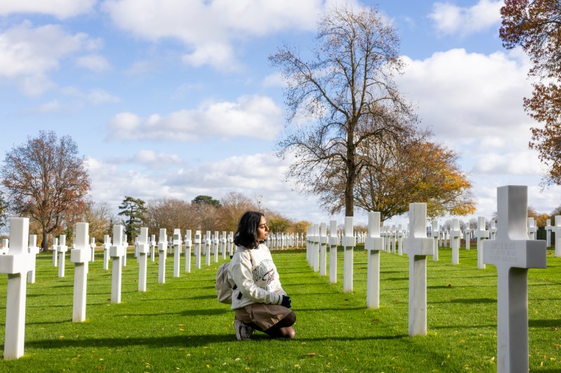 A woman wearing a backpack and a white sweatshirt sits in the middle of a row of cross headstones in a military cemetary.